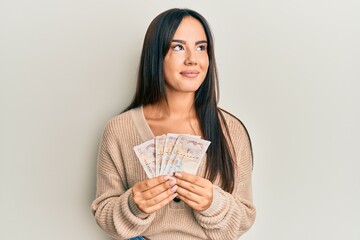 Young beautiful hispanic girl holding 10 united kingdom pounds banknotes smiling looking to the side and staring away thinking.