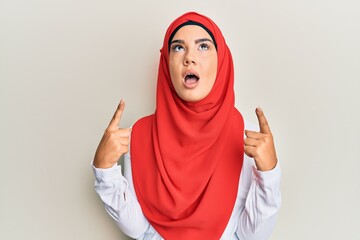 Young beautiful hispanic girl wearing traditional islamic hijab scarf amazed and surprised looking up and pointing with fingers and raised arms.