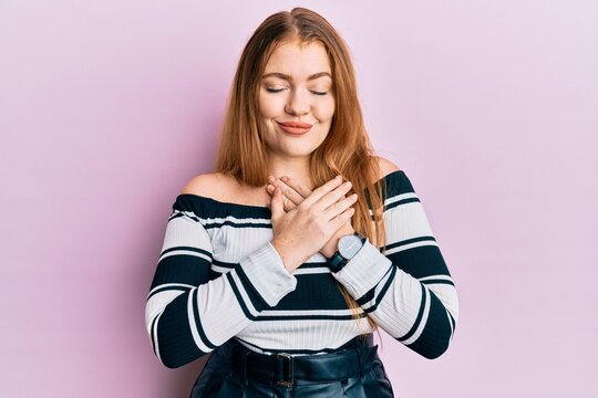 Young beautiful redhead woman wearing striped sweater over pink background smiling with hands on chest, eyes closed with grateful gesture on face. health concept.