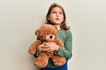 Little beautiful girl hugging teddy bear angry and mad screaming frustrated and furious, shouting with anger looking up.