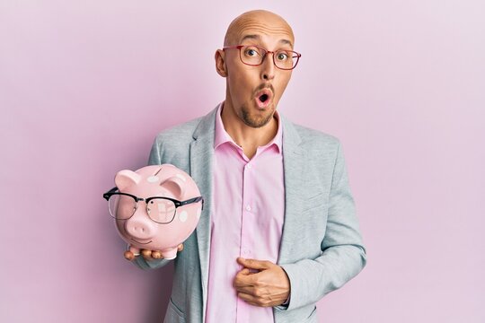 Bald man with beard holding piggy bank with glasses scared and amazed with open mouth for surprise, disbelief face