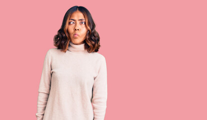 Young beautiful mixed race woman wearing winter turtleneck sweater making fish face with lips, crazy and comical gesture. funny expression.