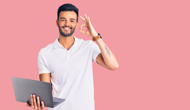 Young handsome hispanic man working using computer laptop doing ok sign with fingers, smiling friendly gesturing excellent symbol