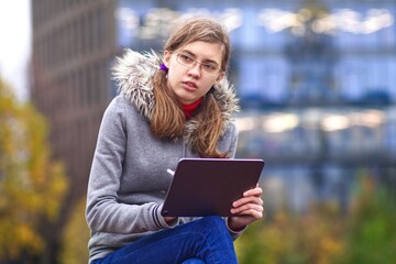 Beautiful young university or college student girl is sitting in the park, drawing on her tablet computer with electronic pen or pencil, studying online with her gadget