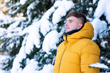 Fototapeta na wymiar Portrait of handsome young man walking in warm yellow down jacket outdoors, guy is breathing deeply fresh air at winter spruce forest in a snowy day
