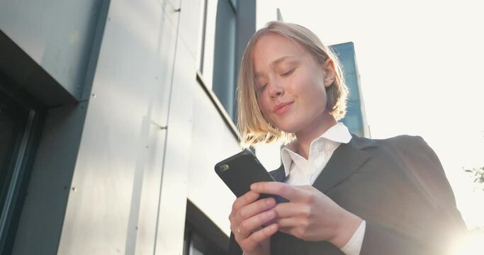 Portrait of Cute Young Female Student texting Messages, walking on warm Sunny Evening. Cute Blonde Girl is browsing her Smarthpone outside, Wind is blowing her stylish Hair. Emotions. Business. Apps.