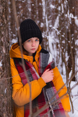 A young girl in warm clothes and with a checkered scarf leans on a tree in a winter forest and looks thoughtfully