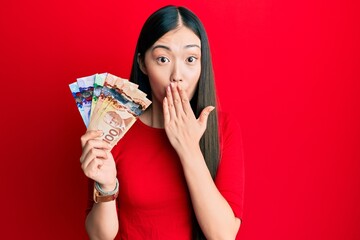 Young chinese woman holding canadian dollars covering mouth with hand, shocked and afraid for mistake. surprised expression