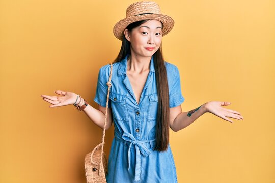 Young chinese woman wearing summer hat clueless and confused expression with arms and hands raised. doubt concept.