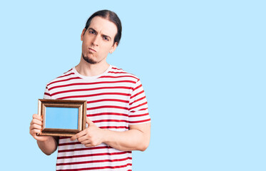Young adult man with long hair holding empty frame thinking attitude and sober expression looking self confident