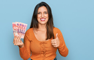 Beautiful hispanic woman holding south african 50 rand banknotes smiling happy and positive, thumb up doing excellent and approval sign