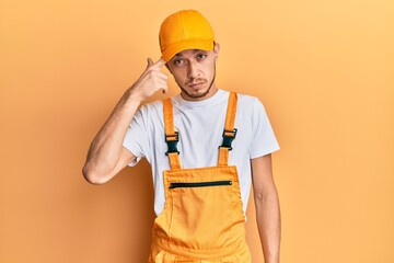 Hispanic young man wearing handyman uniform pointing unhappy to pimple on forehead, ugly infection of blackhead. acne and skin problem