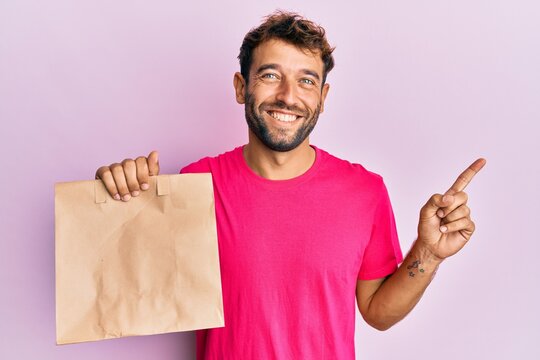 Handsome man with beard holding take away paper bag smiling happy pointing with hand and finger to the side