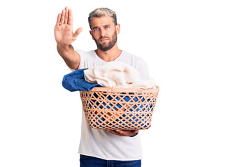 Young handsome blond man holding laundry basket with clothes with open hand doing stop sign with serious and confident expression, defense gesture