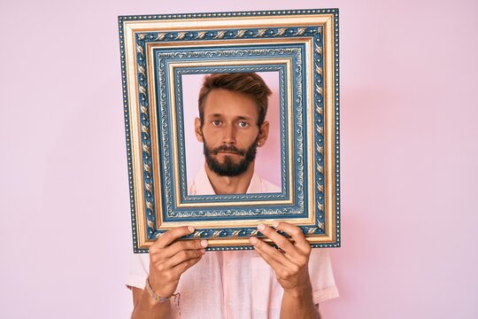 Handsome caucasian man with beard holding empty frame skeptic and nervous, frowning upset because of problem. negative person.