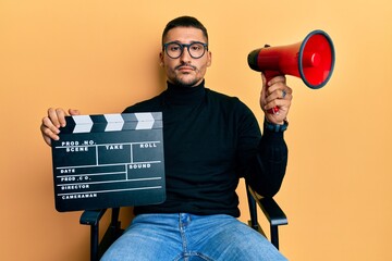 Handsome man with tattoos holding video film clapboard and megaphone relaxed with serious...