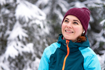 Portrait of happy beautiful young woman smiling in a winter snowy forest, walking in the park and listening to music in wireless earphones