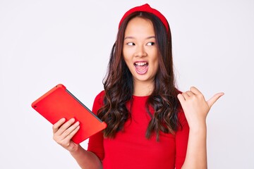 Young beautiful chinese girl holding touchpad pointing thumb up to the side smiling happy with open mouth