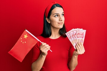 Young hispanic woman holding china flag and yuan banknotes smiling looking to the side and staring away thinking.