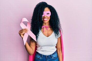 Middle age african american woman wearing super hero costume holding pink cancer ribbon looking...