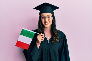 Young hispanic woman wearing graduation uniform holding italy flag looking positive and happy standing and smiling with a confident smile showing teeth