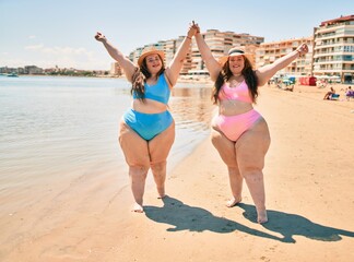 Two plus size overweight sisters twins women happy and proud of their bodies walking at the beach on summer holidays
