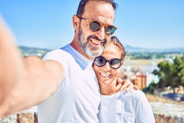 Middle age beautiful couple wearing casual clothes and sunglasses smiling happy. Standing with smile on face making selfie by the camera hugging at town street.