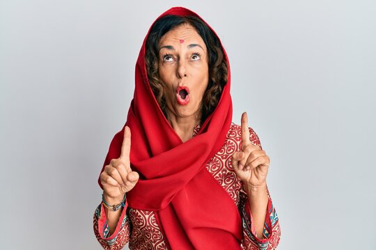 Middle age hispanic woman wearing tradition sherwani saree clothes amazed and surprised looking up and pointing with fingers and raised arms.