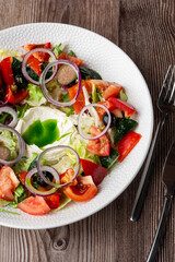 Greek salad with fresh vegetables: tomato, cucumber, red bel pepper, lettuce, onion, olives and cheese. Close-up on a white round plate with cutlery by side on a wooden background. Healthy food.