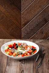A tasty Greek salad with fresh vegetables: tomato, cucumber, red bel pepper, lettuce, onion, olives and cheese. Close-up on a white round plate on a wooden background with a copy space. Healthy food.
