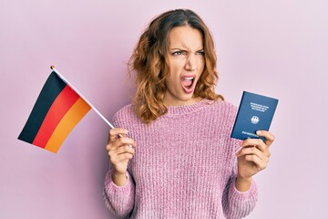 Young caucasian woman holding germany flag and passport angry and mad screaming frustrated and furious, shouting with anger. rage and aggressive concept.