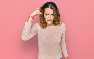 Beautiful young caucasian woman wearing casual sweater angry and mad raising fist frustrated and...