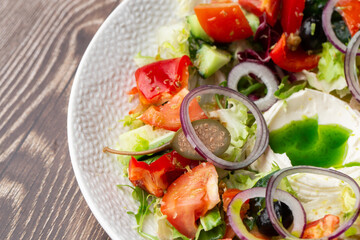 Greek salad with fresh vegetables: tomato, cucumber, red bel pepper, lettuce, red onion, olives and cheese. Macro close-up on a white round plateon a wooden background. Concept of healthy meal.