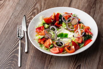Greek salad with fresh vegetables: tomato, cucumber, red bel pepper, lettuce, onion, olives and cheese. Close-up on a white round plate with cutlery by side on a wooden background. Healthy food.