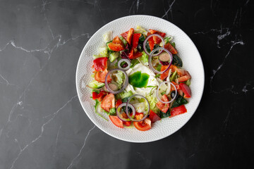 Greek salad with vegetables: tomato, cucumber, red bel pepper, lettuce, onion, olives and cheese. Top view close-up on a white round plate on a black marble background with a copy space. Healthy food.