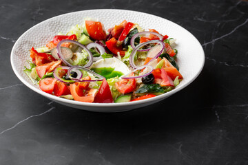 Greek salad with fresh vegetables: tomato, cucumber, red bel pepper, lettuce, onion, olives and cheese. Close-up on a white round plate on a black marble background with a copy space. Healthy food.