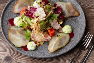 A tasty salad with pear, tomato, mozzarella cheese and fresh lettuce, dressed with a red and green sauces. Close-up on a grey round plate with cutlery by side on a wooden background. Healthy food.