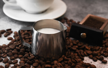 Pitcher with milk surrounded with coffee beans, cup of coffee against concrete background.
