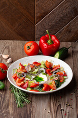 Greek salad with fresh vegetables: tomato, cucumber, red bel pepper, lettuce, onion, olives and cheese. Close-up on a white round plate on a wooden background. Salad menu with ingredients.