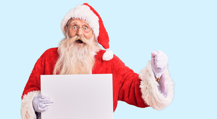 Naklejka premium Old senior man with grey hair and long beard wearing santa claus costume holding banner screaming proud, celebrating victory and success very excited with raised arms