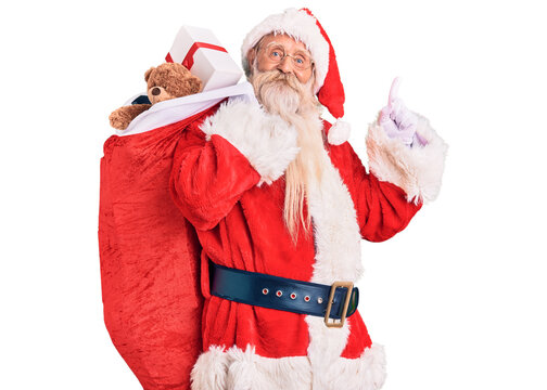 Old senior man with grey hair and long beard wearing santa claus costume holding bag with presents surprised with an idea or question pointing finger with happy face, number one