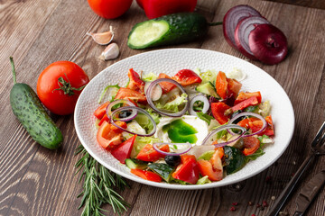 Greek salad with fresh vegetables: tomato, cucumber, red bel pepper, lettuce, onion, olives and cheese. Close-up on a white round plate on a wooden background. Salad menu with ingredients.