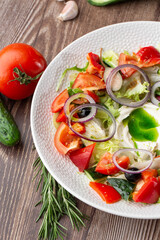 Greek salad with fresh vegetables: tomato, cucumber, red bel pepper, lettuce, onion, olives and cheese. Close-up on a white round plate on a wooden background. Salad menu with ingredients.