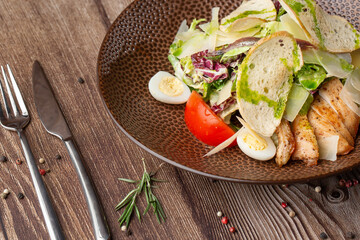 Caesar salad with grilled chicken, tomato, boiled egg, lettuce, croutons dressed with green sauce. Close-up on a brown round plate with spices and cutlery by side on a wooden background.
