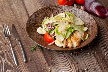 Caesar salad with grilled chicken, tomato, boiled egg, lettuce, croutons and green sauce. Top view close-up on a brown round plate with fresh vegetables and cutlery by side on a wooden background.