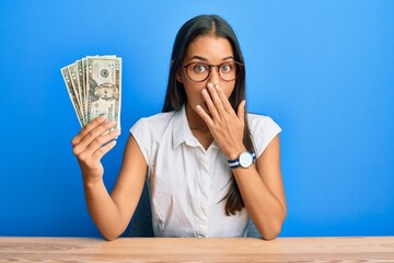 Beautiful hispanic woman holding 20 dollars banknotes covering mouth with hand, shocked and afraid...