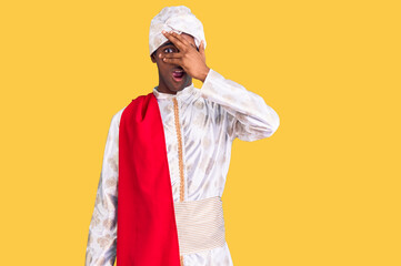 African handsome man wearing tradition sherwani saree clothes peeking in shock covering face and eyes with hand, looking through fingers with embarrassed expression.