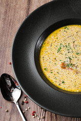 Yellow vegetable soup with onion, tomato, mushrooms, dill and parsley in a black bowl. Macro close-up on a wooden background with a spoon and spices by side. Healthy food menu.