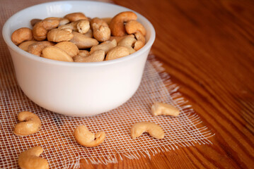 Cashew nuts in a white bowl and five cashews next to a bowl on a wooden table.