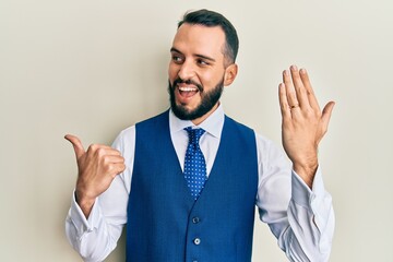 Young man with beard wearing engagement ring pointing thumb up to the side smiling happy with open mouth